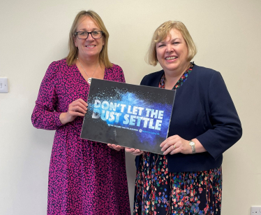 Jane with a representative from Mesothelioma UK holding a 'Don't let the Dust Settle' board